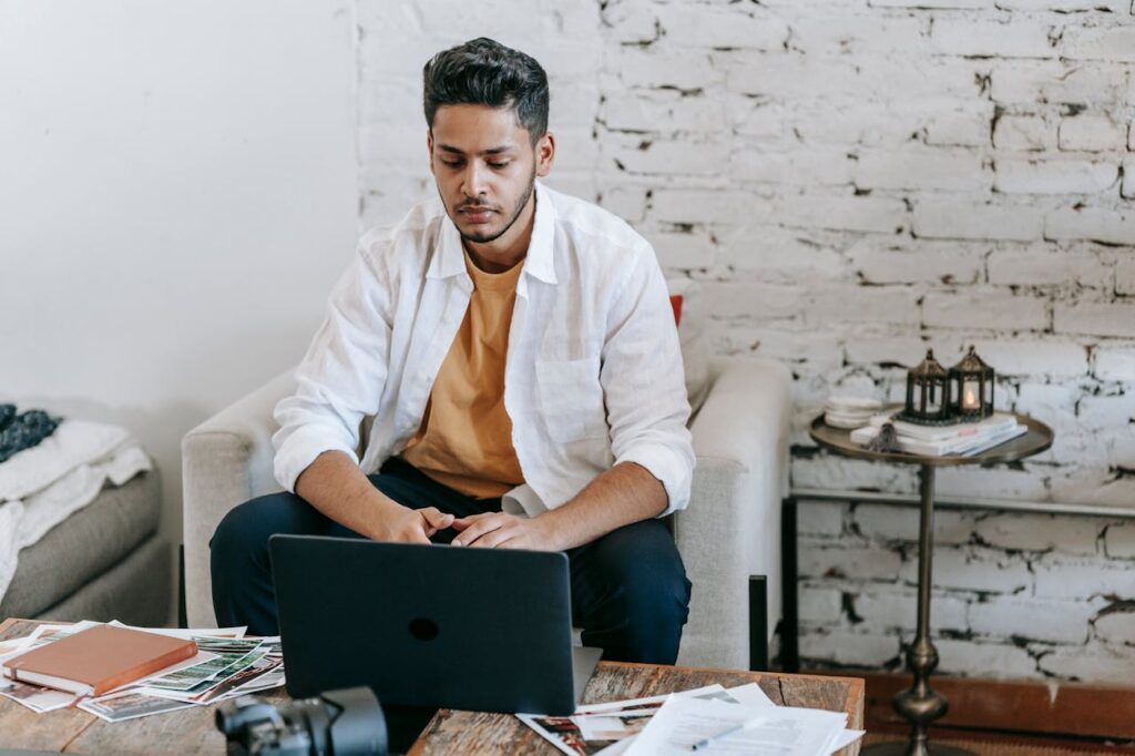 Creative young Hispanic male photographer sitting at table with photo camera and printed photos and working on laptop in modern loft style workplace at home